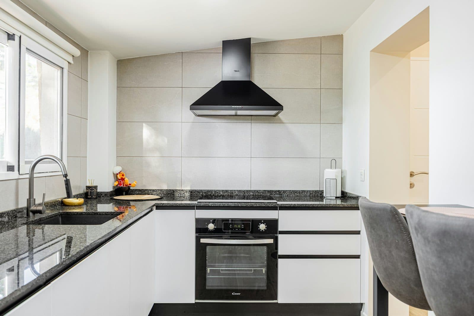 Modern fitted apartment kitchen with white cabinetry, dark stone countertop, and built-in oven beneath a black cooker hood