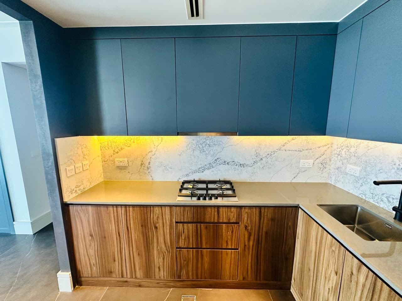 Completed kitchen carpentry with blue upper cabinets, walnut-finish lower units, marble-look backsplash and a built-in gas cooktop.