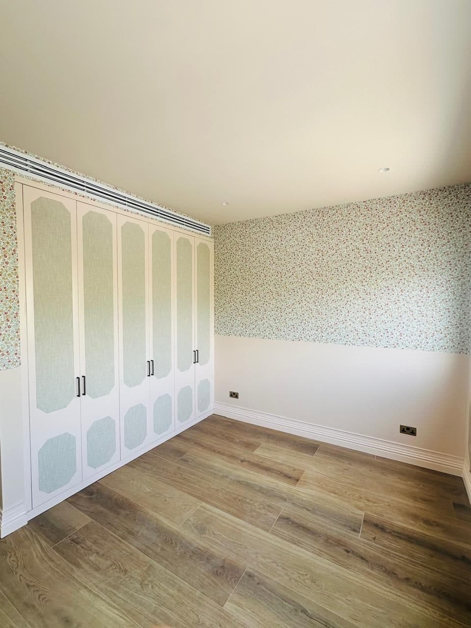 Built-in bedroom wardrobe with pastel, fabric-panelled doors, finished alongside a floral wallpaper accent wall.