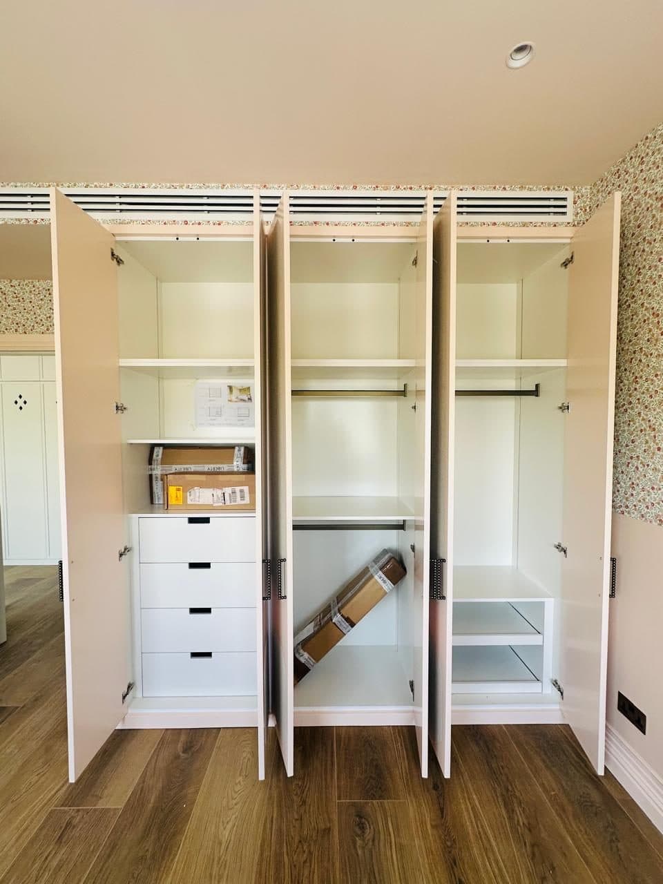 Inside a built-in wardrobe mid-installation, showing the shelving, hanging rails, drawer unit and fold-out shelves being fitted.