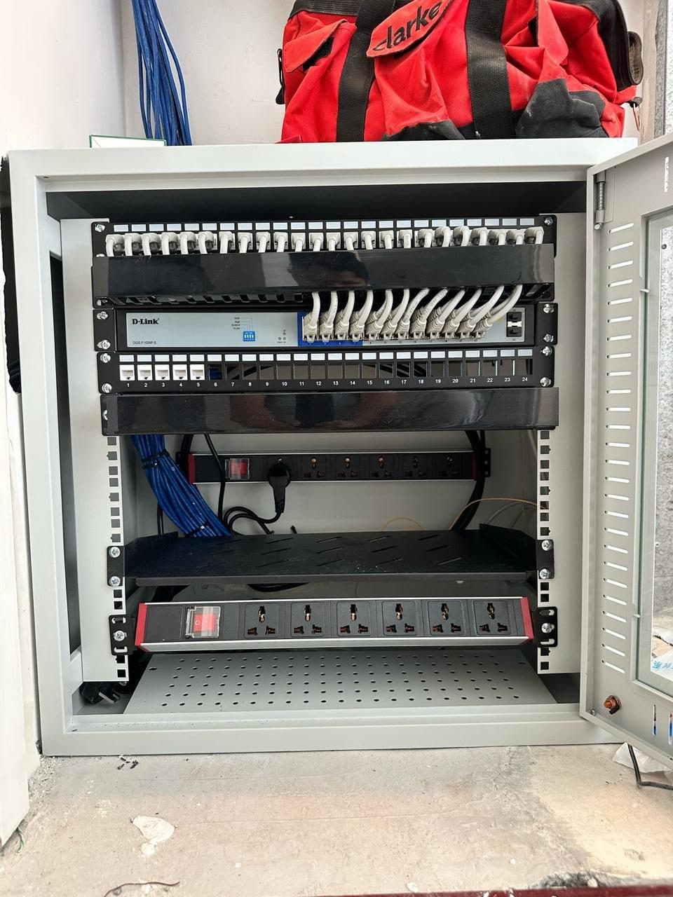Open wall-mounted IT rack showing the network switch, patch panel and power distribution units with cabling terminated.