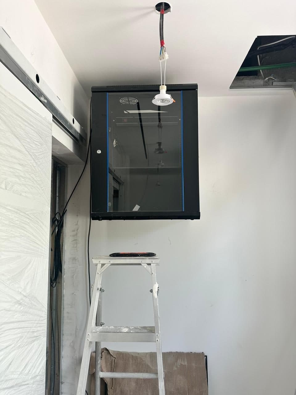 Glass-front network server cabinet installed near the ceiling with cable entry routed through an access panel above.