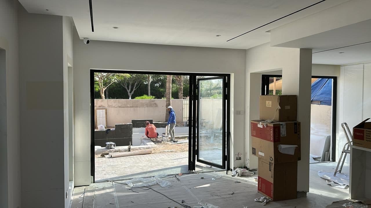 A room mid-construction with bifold glass doors opening to the garden, combining ceiling camera install with exterior masonry and tiling work underway.