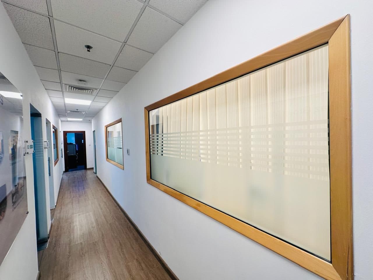 Wood-framed frosted glass panel set into an office corridor wall, finished with vinyl plank flooring and a suspended ceiling.