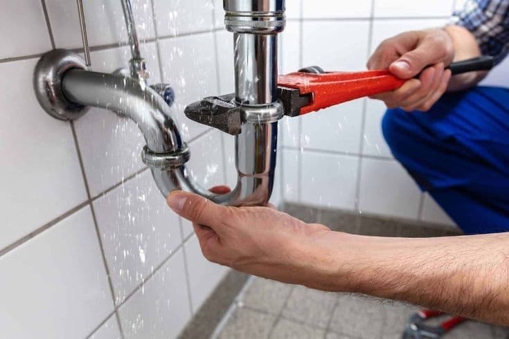 Water spraying from a pipe joint while a plumber repairs it with a wrench